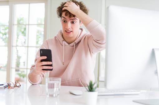 A man is reacting negatively while looking at his phone. He wears a pink sweatshirt and sits at a desk with a computer, mouse, and other desk objects.